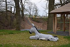 Das Foto zeigt einen Esel aus Holz, der auf einer Wiese neben einem Stall liegt.