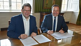 Das Foto zeigt zwei  Männer an eine Tisch, die zwie Dokumente unterschreiben.